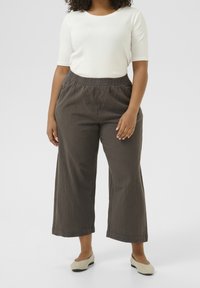 Femme portant un pantalon cropped large marron, un haut blanc à manches courtes et des chaussures plates beiges, se tenant devant un fond uni.