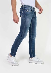Jeans oscuros de corte ajustado hechos de mezclilla con cinco bolsillos, costuras en contraste y herrajes en tono plateado. Llevados con zapatillas blancas.