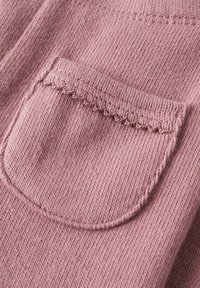 Mīksts rozā adīts audums ar mazu, noapaļotu kabatu, kas rotāta ar viļņveida malas apdari, uz teksturēta džempera.