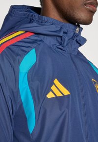 Marineblauw jack met blauwe en rode accenten, ritssluiting aan de voorkant, capuchon en geel logo. Gemaakt van glad, lichtgewicht materiaal, ideaal voor sport.