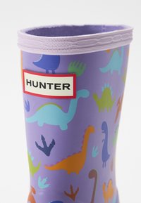Fialové gumové dažďové čižmy s farebnými vzormi dinosaurov, s textúrovaným okrajom a výraznou čiernou a červenou nálepkou "HUNTER".