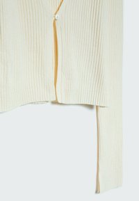 Krémový vroubkovaný pletený cardigan s jedním bílým knoflíkem zapnutým těsně u vrchu a dlouhými rukávy, zobrazený na světlem pozadí.