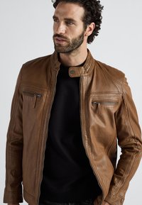Mann mit dunklem lockigem Haar und Bart, der eine braune Lederjacke über einem schwarzen Hemd trägt, leicht zur Seite blickend, mit entspannten Händen.