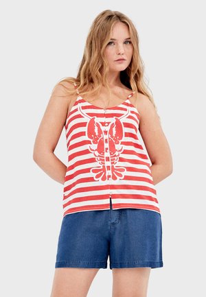Młoda kobieta z długimi blond włosami, ubrana w czerwono-biały top na ramiączkach w paski z czerwonym nadrukiem homara oraz niebieskie dżinsowe szorty.
