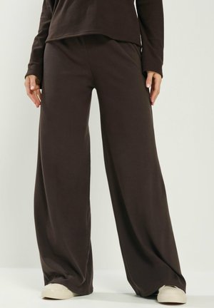 Osoba nosząca luźne, szerokie spodnie w ciemnobrązowym kolorze, dopasowaną bluzkę z długim rękawem oraz białe trampki, stojąca na jednolitym tle.