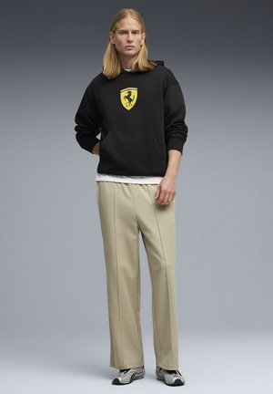 Sweat-shirt à capuche noir avec logo Ferrari, pantalon beige sur mesure, et baskets métalliques. Texture lisse, coupe décontractée, couleurs contrastées.