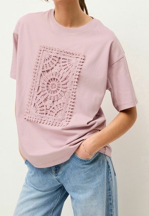 Różowy t-shirt z krótkim rękawem z koronkowym wzorem na środku, o fakturowanym wzorze i luźnym kroju, w zestawieniu z jasnoniebieskimi dżinsami.