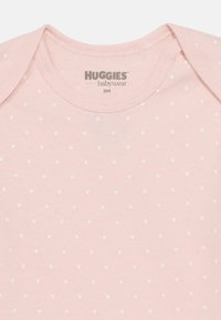Ροζ βαμβακερό κορμάκι μωρού με λευκά πουά, στρογγυλή λαιμ όκοψη και κοντά μανίκια. Η ετικέτα γράφει "HUGGIES babywear 9M."