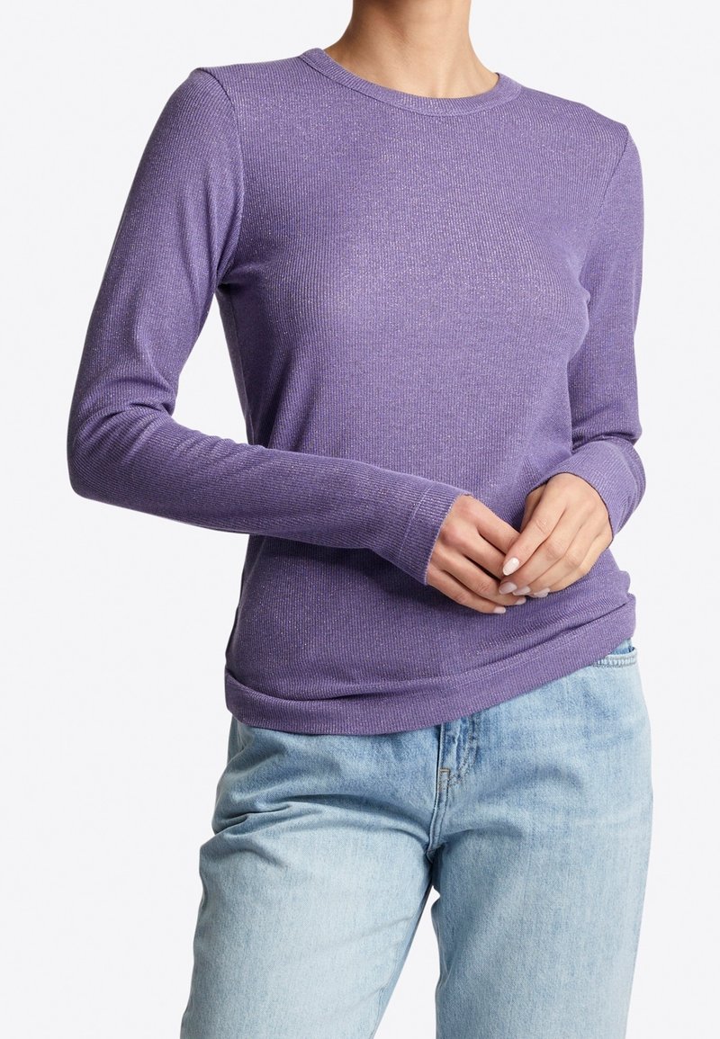 Femme portant un pull violet ajusté à manches longues associé à un jean bleu clair, les mains doucement jointes à la taille.