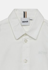 Bela srajca s gumbi iz bombaža, ki ima klasičen ovratnik, prosojne gumbe in belo izvezeno oznako "BOSS" na prsih.