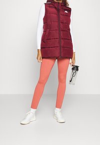 Osoba nosząca bordową kamizelkę puchową, białą bluzkę z długim rękawem, koralowe legginsy, białe sneakersy, trzymająca przezroczystą butelkę wody Nike.