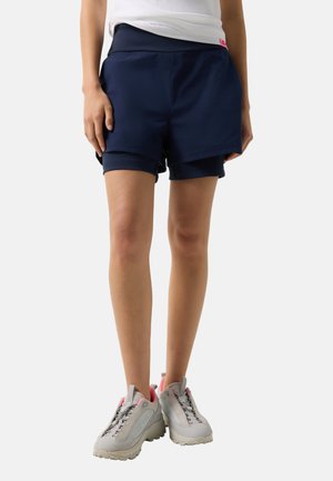 Ben iført marineblå dobbelte shorts og hvide sneakers med pink detaljer, stående mod en hvid baggrund.