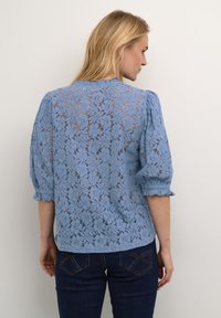 Blusa di pizzo azzurro chiaro con motivo floreale, maniche a sbuffo e polsini arricciati. Il tessuto è trasparente con dettagli delicati.