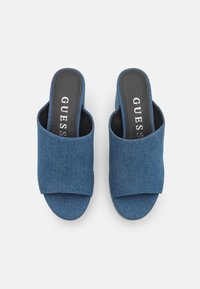 Mule di jeans blu con tacco alto, punta aperta e design con ampia fascia incrociata. La soletta nera presenta il nome del marchio "GUESS" in bianco. Texture morbida.