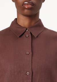 Chemise en lin marron avec un col, présentant trois boutons sur le devant et un tissu légèrement texturé pour un aspect naturel.