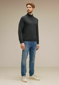 Grå polotröja med en figursydd design, parat med ljusblå slitna jeans och vita sneakers, står mot en neutral bakgrund.