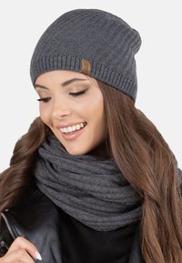 Berretto grigio in maglia con texture a costine e una piccola etichetta in pelle marrone. Indossato con una sciarpa grigia chunky, che mette in risalto uno stile invernale accogliente.