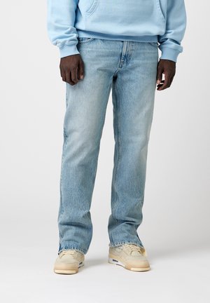 Mężczyzna ubrany w jasnoniebieskie jeansy, beżowe trampki i jasnoniebieską bluzę, stoi na jednolitym tle.