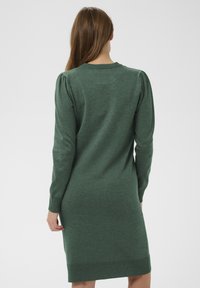 Zaļa adīta kleita ar pieguļošu siluetu, garām piedurknēm un ribotām aprocēm. Mugurpusē ir neliela pūka detaļa plecu šuvēs.