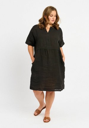 Black linen dress with short sleeves, V-neckline, gathered waist, and side pockets. Smooth texture with a relaxed silhouette. Brown sandals worn.
