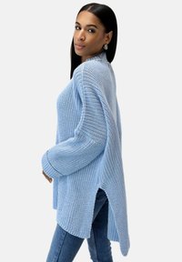Hellblaues oversized Sweatshirt mit strukturiertem Strickmuster, seitlichen Schlitzen und gerippten Bündchen. Kombiniert mit engen Jeans.