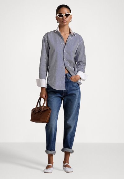 Cămașă cu dungi și guler alb, blugi din denim albastru, pantofi albi slip-on și geantă maro. Modelul poartă ochelari de soare și cercei rotunzi.
