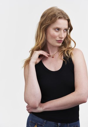Eine rothaarige Frau in einem schwarzen ärmellosen Top und blauen Jeans berührt eine goldene Kette mit einem roten Rosenanhänger und schaut nachdenklich zur Seite.