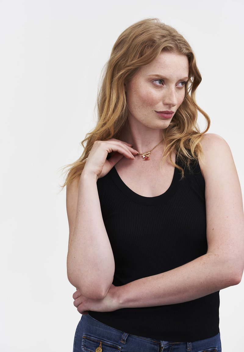 Eine rothaarige Frau in einem schwarzen ärmellosen Top und blauen Jeans berührt eine goldene Kette mit einem roten Rosenanhänger und schaut nachdenklich zur Seite.