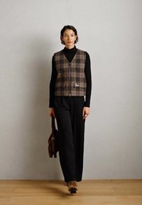 Žena v kostkované vestě na zip přes černý rolák a kalhoty, držící hnědou tašku, stojící na dřevěné podlaze proti jednoduché zdi.
