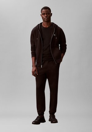 Jauns vīrietis stāv, valkājot melnu rāvējslēdzēja kapuci, melnu T-kreklu, melnas sporta bikses un melnas sporta kurpes vienkāršā pelēkā fonā.