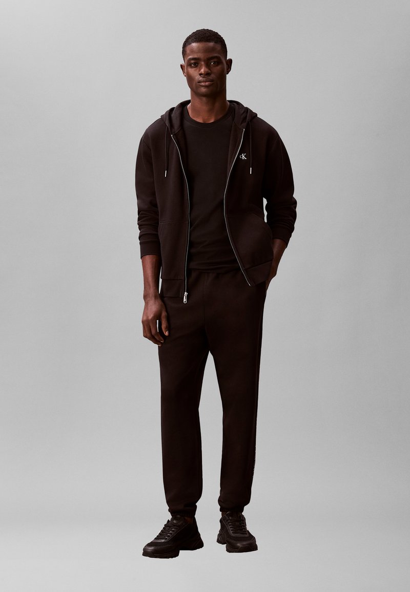 Νέος άνδρας που στέκεται, φορώντας μαύρο φούτερ με φερμουάρ, μαύρο μπλουζάκι, μαύρο παντελόνι φόρμας και μαύρα αθλητικά παπούτσια, μπροστά σε απλό γκρι φόντο.