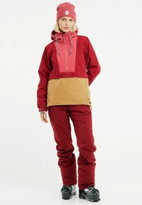 Rode skijas met een roze paneel, tan onderzijde, trekkoordcapuchon en contrasterende stiksels, gecombineerd met bijpassende rode skibroeken en zwarte laarzen.