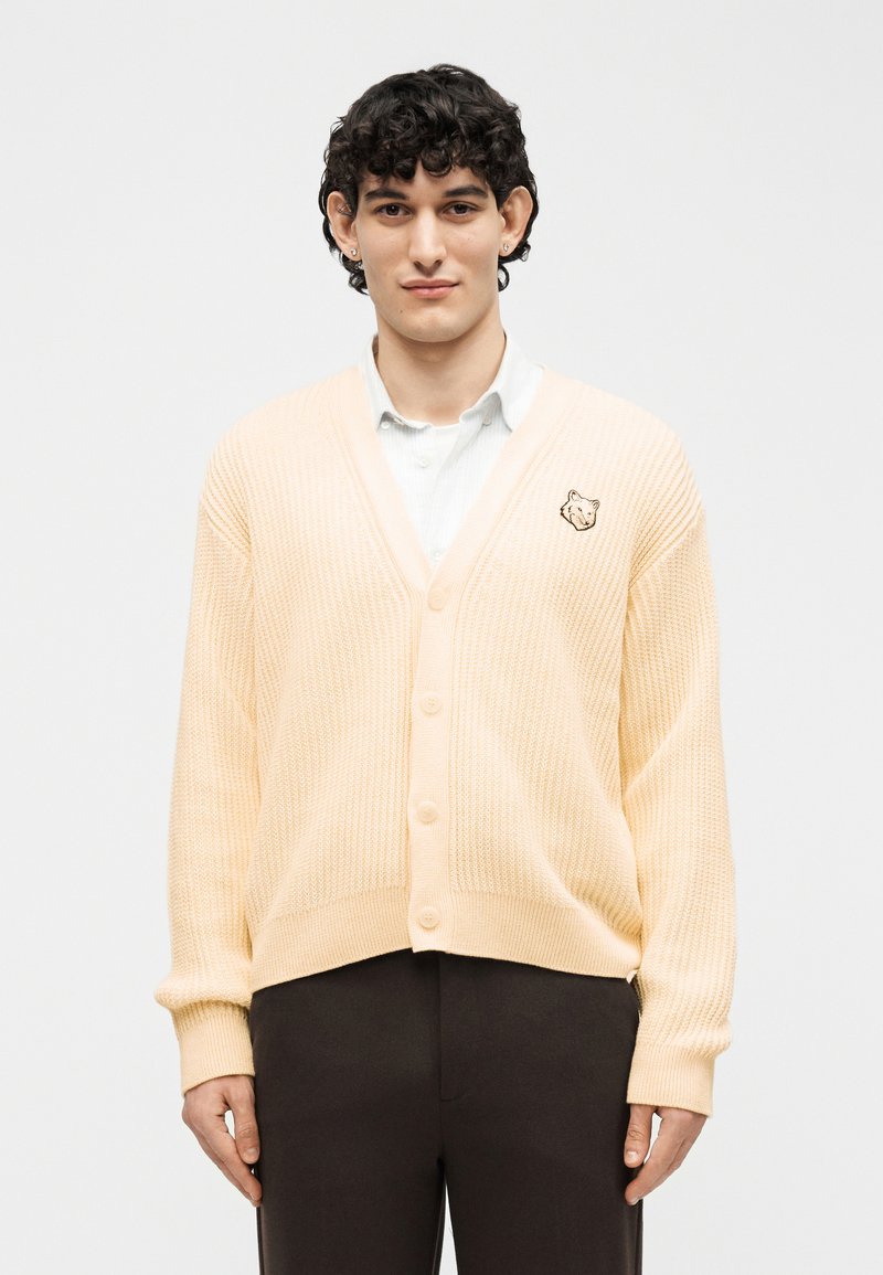 Νεαρός άνδρας με σκούρα σγουρά μαλλιά, φοράει κρεμ ζακέτα με κουμπιά και ένα μπαλώμα με κεφάλι αλεπούς πάνω από ένα λευκό πουκάμισο με γιακά.