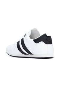 Zapatilla deportiva blanca con acentos en negro a rayas, material de cuero texturizado, punta redondeada y una pestaña negra en el talón con un logo.