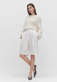 Krämfärgad stickad tröja med löst sittande ärmar, parat med högmidjade, ljusbeige, veckade shorts. Skor i spetsig modell med ormskinnsmönster.