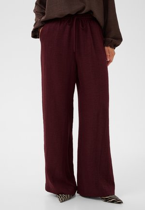 Femme portant un pantalon ample bordeaux foncé avec taille à cordon et des chaussures pointues zébrées, debout contre un fond uni.