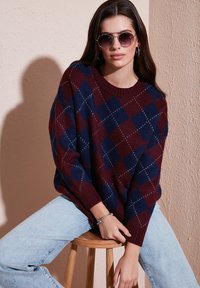 Pull en tricot de couleur bordeaux et marine avec un motif en losange, coupe décontractée, col et poignets côtelés ; associé à un jean bleu clair et des lunettes de soleil.