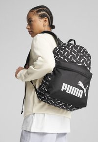 Juodas kuprinės, su balta Puma logo ir pakartotiniais katės raštais, turi reguliuojamus diržus ir užtrauktuką priekinėje kišenėje.