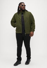 Oliven grøn bomberjakke med høj krave, lynlåslukning og ribbede manchetter, båret over en sort skjorte og kombineret med sorte jeans og sneakers.