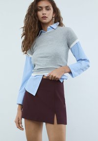 Giovane donna con lunghi capelli mossi che indossa orecchini a stella, una camicia a righe blu con un maglione grigio a maniche corte e una gonna bordeaux con uno spacco frontale.