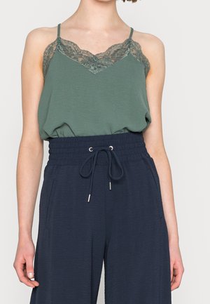 Femme portant un caraco vert bordé de dentelle rentré dans un pantalon taille haute bleu marine à cordon avec poches, debout devant un fond uni.