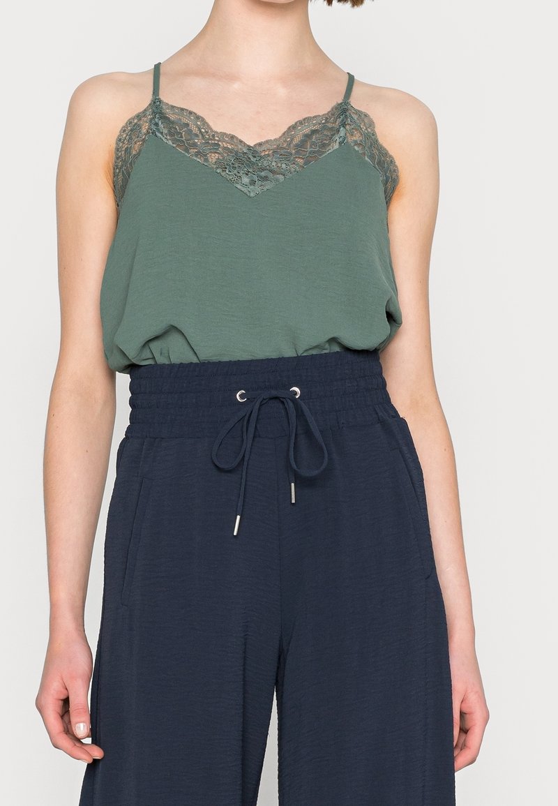 Femme portant un caraco vert bordé de dentelle rentré dans un pantalon taille haute bleu marine à cordon avec poches, debout devant un fond uni.