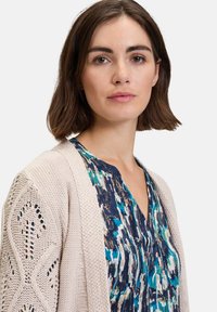 Lichtbeige gebreide cardigan met opengewerkte patronen op de mouwen, gecombineerd met een blauw en groen abstract patroon top met een v-hals.