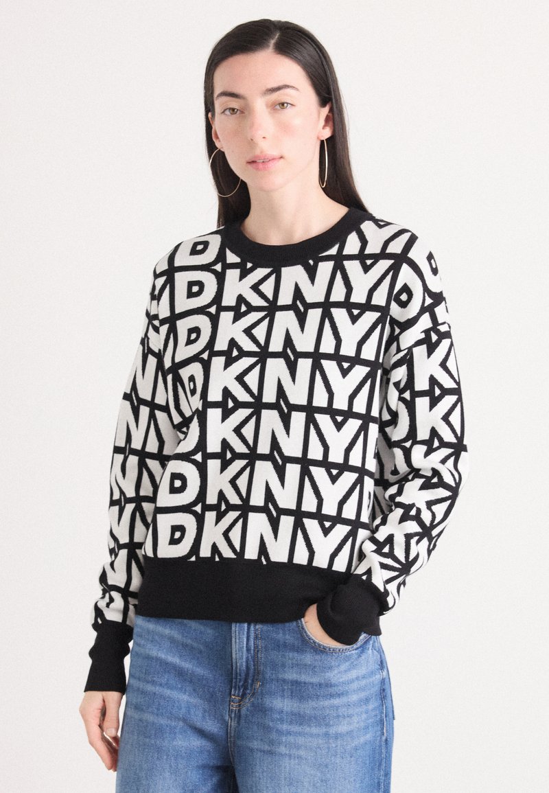Juodai balta DKNY megztinis, turintis ryškią kartotinę tekstų raštą, apvalią kaklo iškirptę ir raibotus rankogalius bei apatinę dalį. Dėvimas su mėlynomis džinsais.