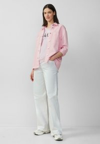 Chemise rose clair à boutons sur un T-shirt blanc, accompagnée de pantalons larges blancs et de baskets gris clair. Design simple et décontracté.