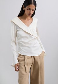 Blusa crema con escote en V y cuello drapeado, mangas largas y cintura ajustada. Combinada con pantalones beige de cintura alta, con pliegues.