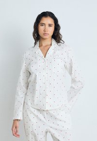 Camicia da pigiama bianca a maniche lunghe con piccoli motivi a cuore rossi, bottone centrale e taschino sul petto. Tessuto morbido e leggero con una vestibilità comoda.