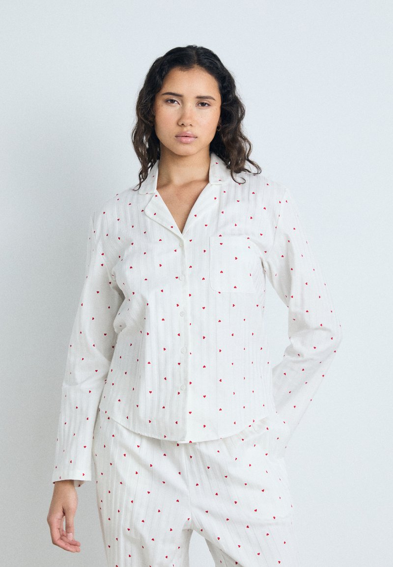 Camicia da pigiama bianca a maniche lunghe con piccoli motivi a cuore rossi, bottone centrale e taschino sul petto. Tessuto morbido e leggero con una vestibilità comoda.