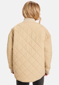 Beige quiltet jakke med høyt krage, som har et diamantmønster og ribbestrikkede mansjetter. Designet er løstsittende og strømlinjeformet.