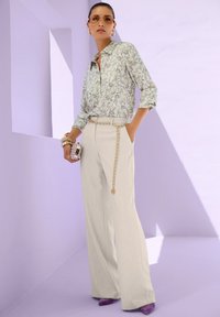 Vrouw in een paisley blouse, hoog-wasbeige wijde broek met een gouden kettingriem, paarse hakken, zonnebril en een kleine versierde clutch in de hand.